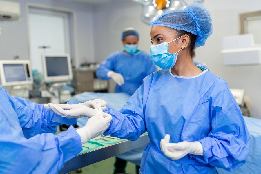 Young nurse in protective mask and workwear helping surgeon with gloves while both preparing for operation